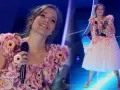 Zuzanna Irena Jurczak w kwiecistej sukni na scenie, trzyma mikrofon i uśmiecha się do publiczności.