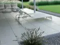 Nowoczesny taras betonowy z leżakami i sofą, otoczony trawą i kamieniami. Idealne miejsce na relaks.