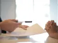 Dłoń z długopisem nad dokumentem, druga dłoń złożona w geście prośby lub oczekiwania. Wszystko dla zgodności z oryginałem.