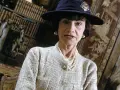 Iconica stilista Coco Chanel, con cappello blu e perle, in un ambiente elegante con libri e decorazioni.