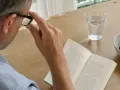Un uomo con occhiali legge un libro, forse perché vede a righe. Sul tavolo, un bicchiere d'acqua e una ciotola di prugne.