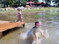 Dwie dziewczynki pluskają się w wodzie przy drewnianym pomoście. W tle plażowicze i drzewa. Wakacje nad jeziora lubelskie.