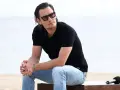 Bartosz Gelner w okularach przeciwsłonecznych i czarnej koszulce polo siedzi na plaży, oparty o drewnianą ławkę.
