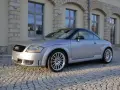 Silberner Audi TT 2005 mit schwarzem Dach parkt auf Kopfsteinpflaster vor einem Gebäude mit Steinbögen.