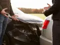 Dwie osoby dyskutują przy uszkodzonych samochodach po kolizji. Mężczyzna gestykuluje, wskazując na zniszczony przód czarnego auta. Kobieta stoi przy srebrnym samochodzie, również wyrażając swoje emocje. Trudno ustalić numer polisy OC bez dokumentów.