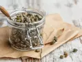 Barattolo di vetro pieno di capperi sotto sale, con un cucchiaino di legno. Alcuni capperi sparsi sul foglio di carta marrone.