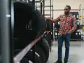 Uomo in un negozio di pneumatici, esamina gomme ricostruite come riconoscerle. Scaffali pieni di pneumatici.