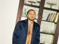 Hombre con pelo rapado y barba, viste una chaqueta abierta sobre el pecho. Detrás, una estantería con libros y vasos.