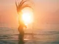 Silhouette d'une femme jetant ses cheveux dans l'eau au coucher du soleil, avec un effet de lens flare photoshop.