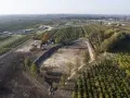 Trwa budowa stawu rybnego. Widać koparki, maszyny budowlane i przygotowany teren pod zbiornik wodny, otoczony sadem i autostradą.