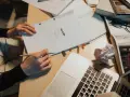 Manos escribiendo un guion cortometraje, rodeado de libros, gafas y un portátil.