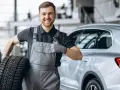 Meccanico sorridente con una gomma primo equipaggiamento in mano, fa il pollice in su accanto a un'auto.