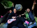 Energiczna ekipa w kosmicznej scenografii, grająca polski folk rock. Kobieta z różową gitarą, mężczyzna ze skateboardem, wesołe miny i podniesione ręce.