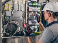 Elektryk w kasku przeprowadza przegląd instalacji elektrycznej, używając multimetru do pomiarów.