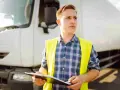Młody mężczyzna w kamizelce odblaskowej trzyma dokumenty przy białej ciężarówce, gotowy do rozpoczęcia transportu.