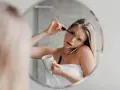 Une femme, enroulée dans une serviette, applique son mascara tout en tenant une tasse et en parlant au téléphone. Le mascara qui coule sur son visage témoigne de sa matinée mouvementée.
