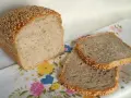 Chleb z kaszą gryczaną, posypany sezamem, leży obok dwóch kromek na kwiecistym obrusie.