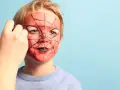 Ein Kind wird mit einem Spiderman-Gesicht bemalt. Rote Farbe und schwarze Spinnennetzmuster verwandeln das Gesicht in eine Superheldenmaske.