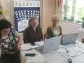 Trzy kobiety pracują przy stołach w sali, która wygląda jak szkoła specjalna. Na ścianie wisi tablica z motylami i napisem "Zgadula".
