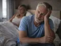 Un homme âgé, le visage pensif, se demande pourquoi ma libido est en berne, tandis qu'une femme est assise dans le lit derrière lui.