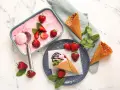 Ein Behälter mit rosa Obst Eis, Erdbeeren und Minze. Ein Eisbecher mit Eis und Schokoladensauce.