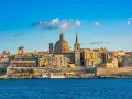 Malta, wyspa do kogo należy? Widok na Vallettę z kopułą i wieżą kościelną, otoczoną zabytkową architekturą, nad błękitnym morzem.