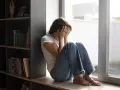Kobieta siedzi przy oknie, zakrywając twarz dłońmi. Wokół panuje nastrój melancholii, odzwierciedlający dystymię.