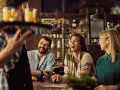 Un camarero sirve bebidas a un grupo de amigos riendo en un bar. La conversación animada sugiere que aún no es tarde para el ruido.
