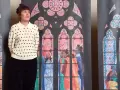 Une femme pose devant des vitraux stylisés, évoquant les vitraux de Notre Dame. Les scènes colorées représentent des figures humaines et des motifs floraux.