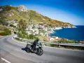 Un motociclista esplora le curve panoramiche dell'Elba, con il mare blu e un villaggio pittoresco sullo sfondo.