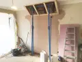 Travaux de rénovation : des étais soutiennent une structure en bois au-dessus d'une zone où l'on s'apprête à casser une cheminée. Matériaux de construction et outils au sol.