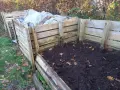Drewniany kompostownik w ziemi, częściowo przykryty plandeką. Widać jesienne liście i trawę.