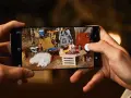 Une photo de téléphone capture un chien blanc endormi lors d'un camping. L'écran du téléphone montre une scène de camping avec une tente, une chaise pliante et un feu de camp.