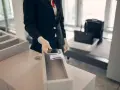 Pracownik lotniska w rękawiczkach kładzie telefon i karty pokładowe do pojemnika. W tle widać bagaż na taśmie. Zapalniczka w samolocie jest zabroniona.