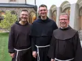 Drei Franziskaner Minoriten in braunen und schwarzen Kutten lächeln in die Kamera vor einer Klosterfassade.