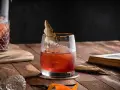 Czerwony koktajl z lodem i liściem laurowym w szkle, obok karafki i starej książki. To może być drink z wermutem.