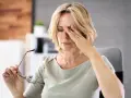 Kobieta z blond włosami masuje czoło, cierpiąc na ból z tyłu oka. W dłoni trzyma okulary.