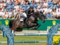 Cavalier en plein saut d'obstacles, le cheval est en l'air, le cavalier porte un casque noir. Le public est en arrière-plan.