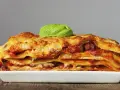 Una porción generosa de lasaña de carne, con capas de pasta, salsa y queso dorado, coronada con una hoja de albahaca fresca.