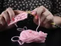 Händerna stickar en rosa v-ringning med ljusa trästickor. En garnnystan ligger bredvid.