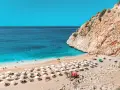 Piękna plaża z turkusową wodą i skalistym klifem. Ludzie odpoczywają na leżakach pod parasolami. Już widać, za ile są wakacje!