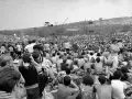 Tysiące ludzi siedzi na trawie, patrząc na scenę festiwalu. To jest Woodstock, gdzie jest muzyka i wolność.