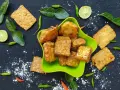 Knusprig gebratener Tempeh, ein fermentiertes Sojaprodukt, liegt in einer grünen Schale. Dazu garnieren Limettenscheiben, grüne Chilis und Blätter.