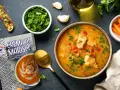 Un tazón de sopa de pescado casera con trozos de pescado, tomates y perejil, junto a una tostada, zanahorias y un paquete de crema de marisco.