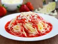 Ein Teller mit Spaghetti Eis, garniert mit Erdbeersauce und weißen Schokostückchen. Frische Erdbeeren und geriebene Schokolade im Hintergrund.