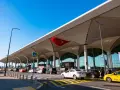 Nowoczesny budynek lotniska Bodrum z charakterystycznym zadaszeniem. Przed terminalem ruch samochodowy, w tym żółte taksówki.