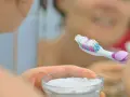 Ręka trzymająca szczoteczkę do zębów z białym proszkiem, obok miseczka z sodą oczyszczoną. Domowa pasta do zębów gotowa.