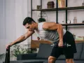 Mężczyzna wykonuje trening obwodowy w domu, podnosząc hantle. Oparty o krzesło, skupiony na ćwiczeniu.