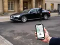 Hand hält Smartphone mit Uber-App, die eine Fahrt mit uber premium cars anzeigt. Ein Fahrer steht neben einem schwarzen Audi.