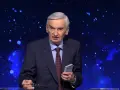 Stars twinkle behind a man in a suit, holding glasses and a small book. He looks like he's been on "Jeden z dziesięciu" for a long time.